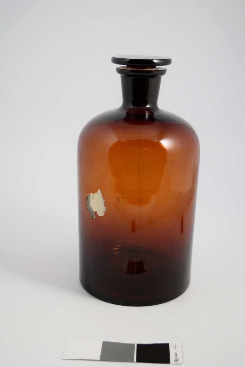 Brun glassflaske, smal hals, brun glasspropp. Brukt til oppbevaring av løsninger eller løsemidler.