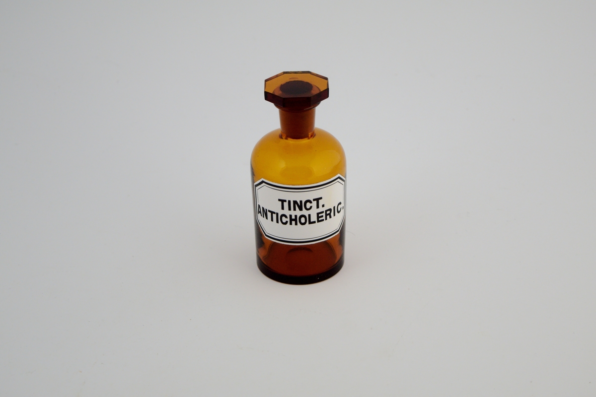 Brun glasskrukke med brun glasspropp. Hvit etikett med sort skrift. Tinct. anticholerica ble brukt som et beroligende middel, spesielt på 1800-tallet.