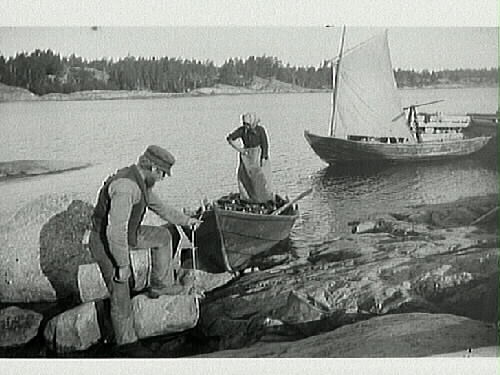 Ett fiskarpar, kvinnan står i båten och mannen håller in ekan mot stenhällarna. Även en segelskuta syns i bild.