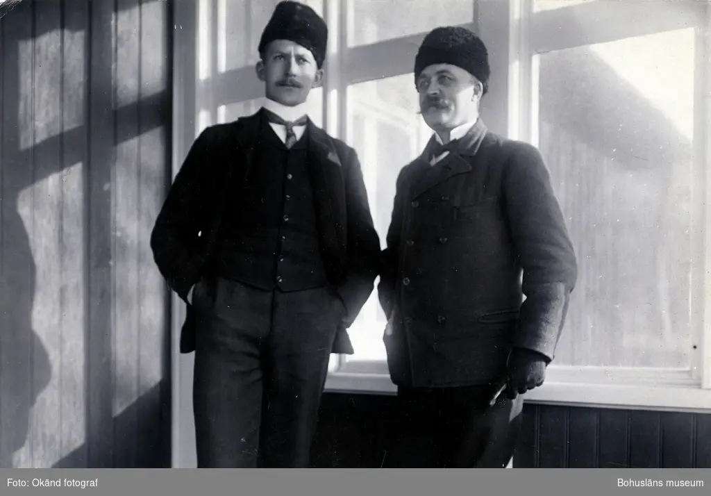 Två män i pälsmössa och kostym står framför ett fönster