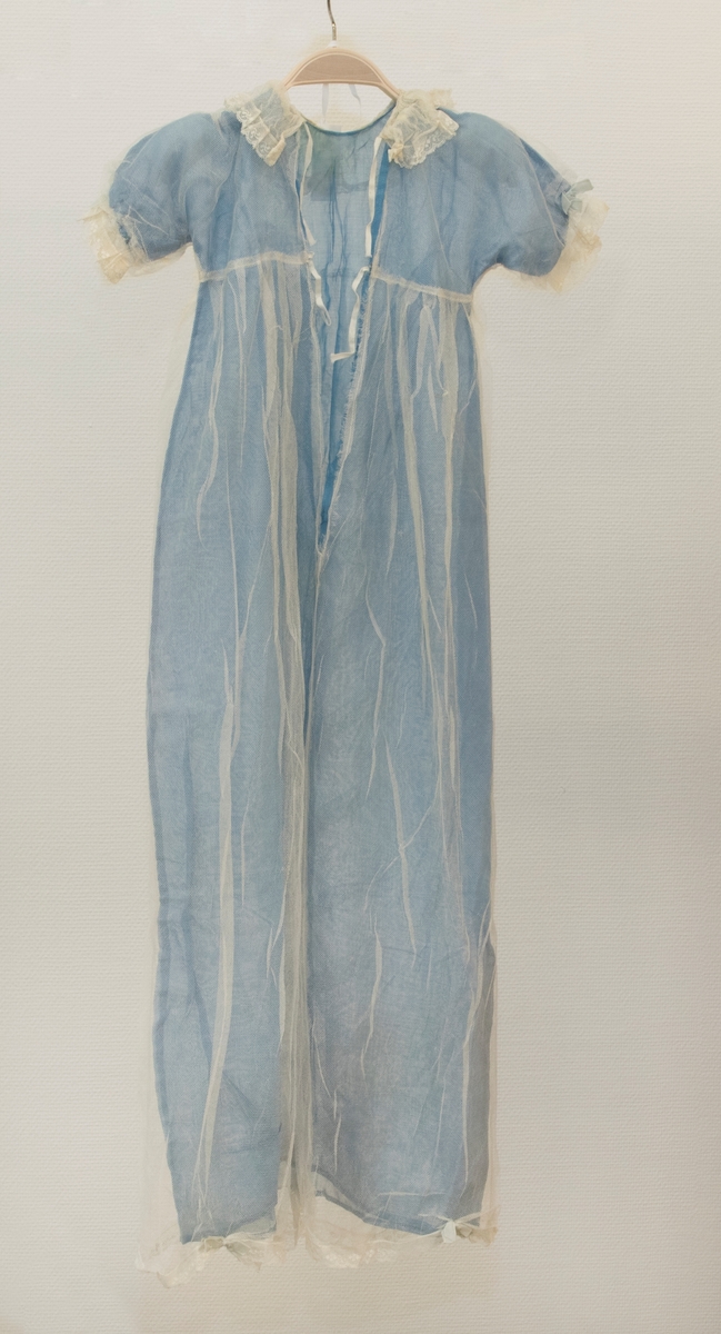 Doplänning bestående av överklänning av vit tyll, och underklänning av ljusblått tunt tyg vävt i tuskaft. Klänningen har hög midja (under armhålorna), vid underdel (tyllklänningen är vidare än underklänningen), korta ärmar och öppning i ryggen, som knyts ihop med två par vita bomullsband. Halsringningen, ärmsluten och klänningens nederkant är prydda med brodyr. Samma typ av brodyr dekorerar klänningens mittparti framtill, tillsammans med ljusblåa rosetter av sidenband, och löst hängande sidenband från midjan.