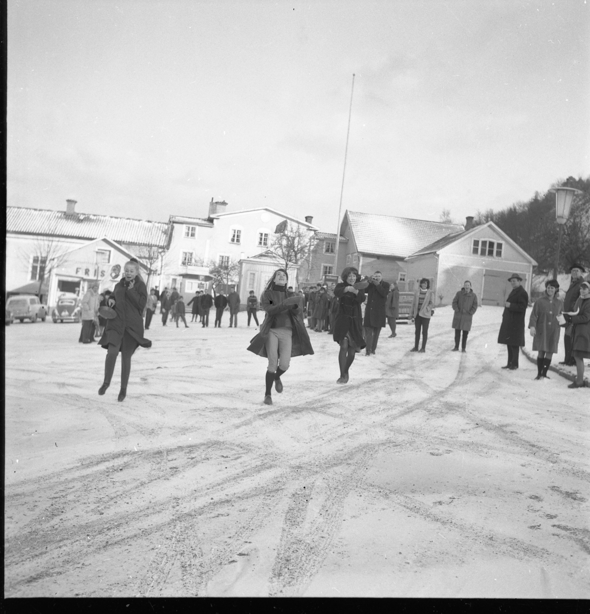 Grännaskolan arrangerar "Pannkakslopp". Tre flickor springer över ett snötäckt Gränna torg, från norra delen mot den södra, med en stekpanna med pannkaka i. Från vänster Marita Wallmo, Susanna Wollin samt okänd till höger. I bakgrunden kvarteret Ångbåten med bl a Risbergs frisörsalong och bakom den hade Aston Bäck en cykelverkstad.