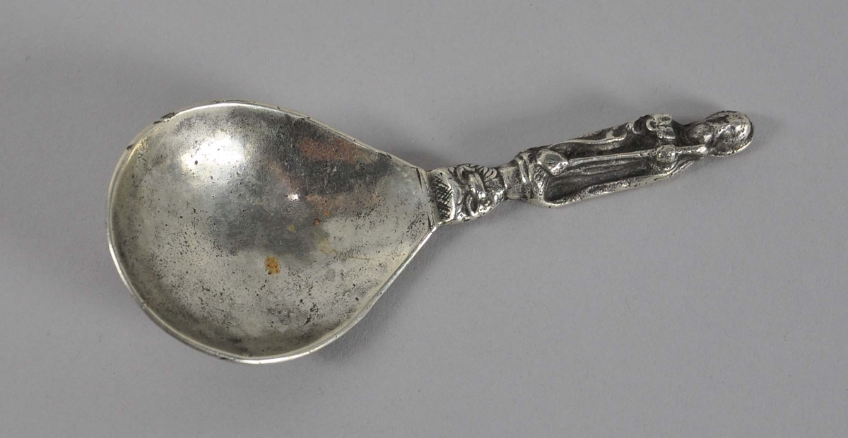 Rikt dekorert skje, av sølv. Skaftet har ikonisk motiv av mann med stav og bok. På baksiden av skjehodet er det inngravert innskrift rundt en mann med trompet og ball.