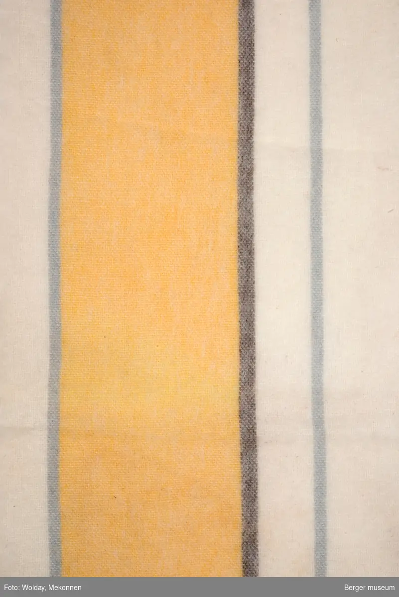 En avlang pleddprøve med stripemønster langs lengderetning; et bredt gult felt er adskilt av grå striper på hver side, og har hhv to og ett stripefelt i offwhite på hver side. Den ene lyse stripefeltet er delt av en smal grå stripe. Prøven har klippede kanter, og frynsekant på ene kortenden.