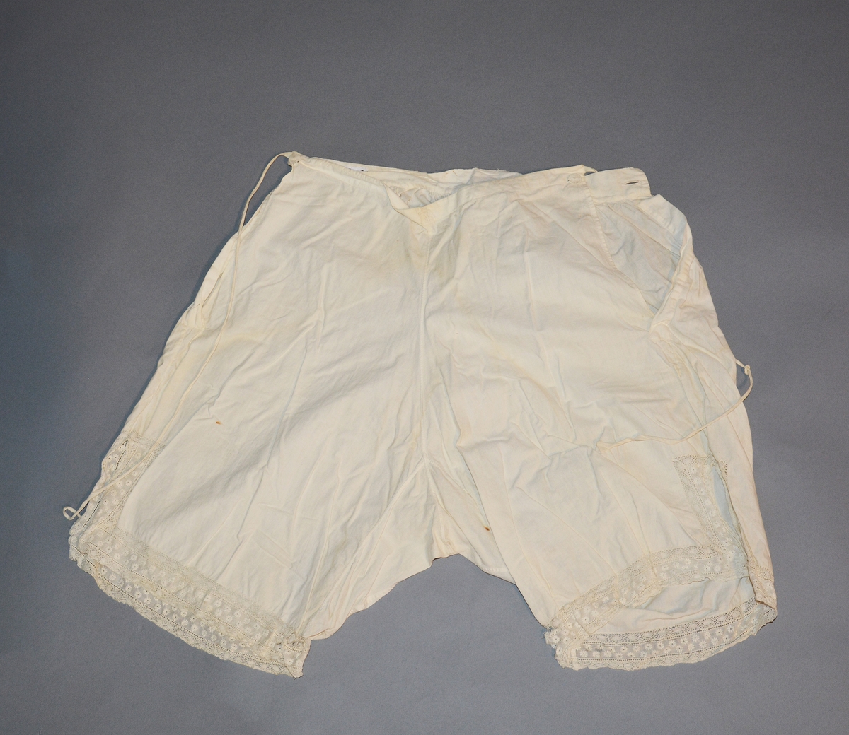 Underbukse for dame i bomullslerret. Åpning i sidene med knytebånd, knapp og knapphull. Bred linning bak, linning foran skåret ned i spiss og kantet med bendelbånd. Innfelt bomullsblonde nede på bena, og kantet med bomullsblonde.