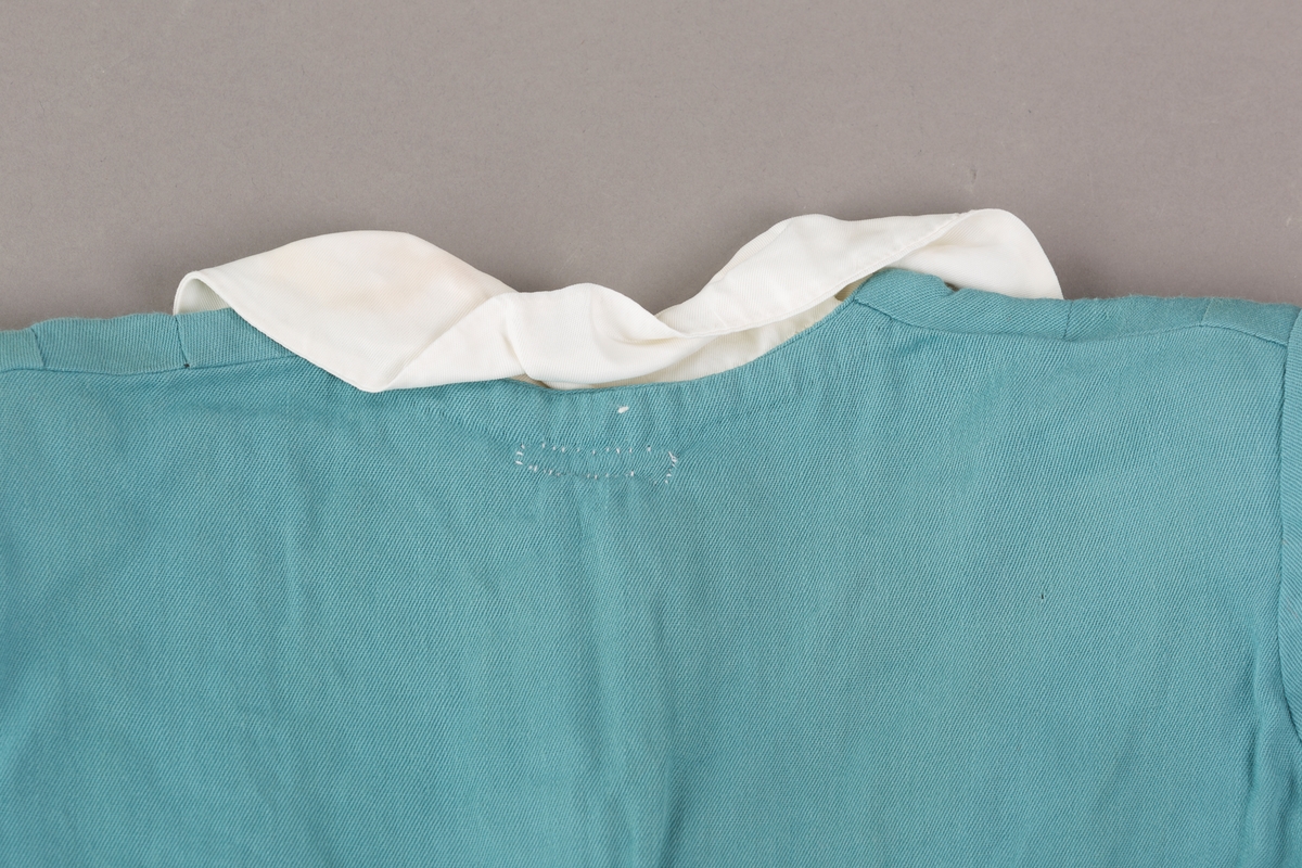 Form: Innsvingt liv, korte ermer, plissert skjørt, fastsydd belte.
Krave.
Bærestykket har vertikale stolper på hver side fra skulder ned til liv, med sydde legg.
Åpning foran med trykknapper. 3 pynteknapper utenpå.