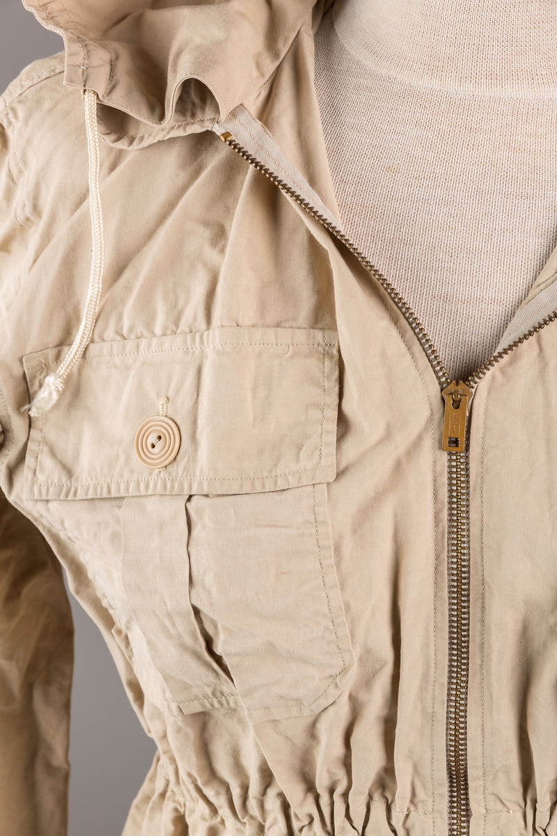 Form: Vanlig anorakkfasong med strikk i livet. 2 brystlommer med knapp.
Hette med glidelås på topp midt, snøring.
Glidelås helt ned foran.
Innermansjett med strikk i ermene.