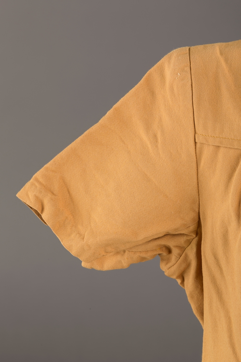 Form: Korte ermer, livet rynket til bærestykke foran, plissert skjørt, tre pynteknapper, en trykknapp, forlomme.