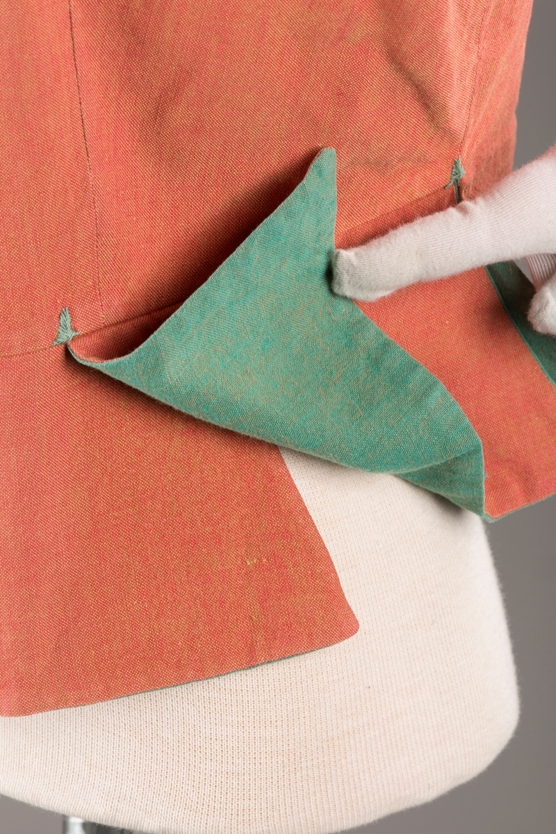 Rustrødt liv med grønn kant og fór, seks skjøter. Håndsydd forsterkning i kontrastfarge øverst i skjøtesplittene. Lukkes med tinnhekte og silkesnor.