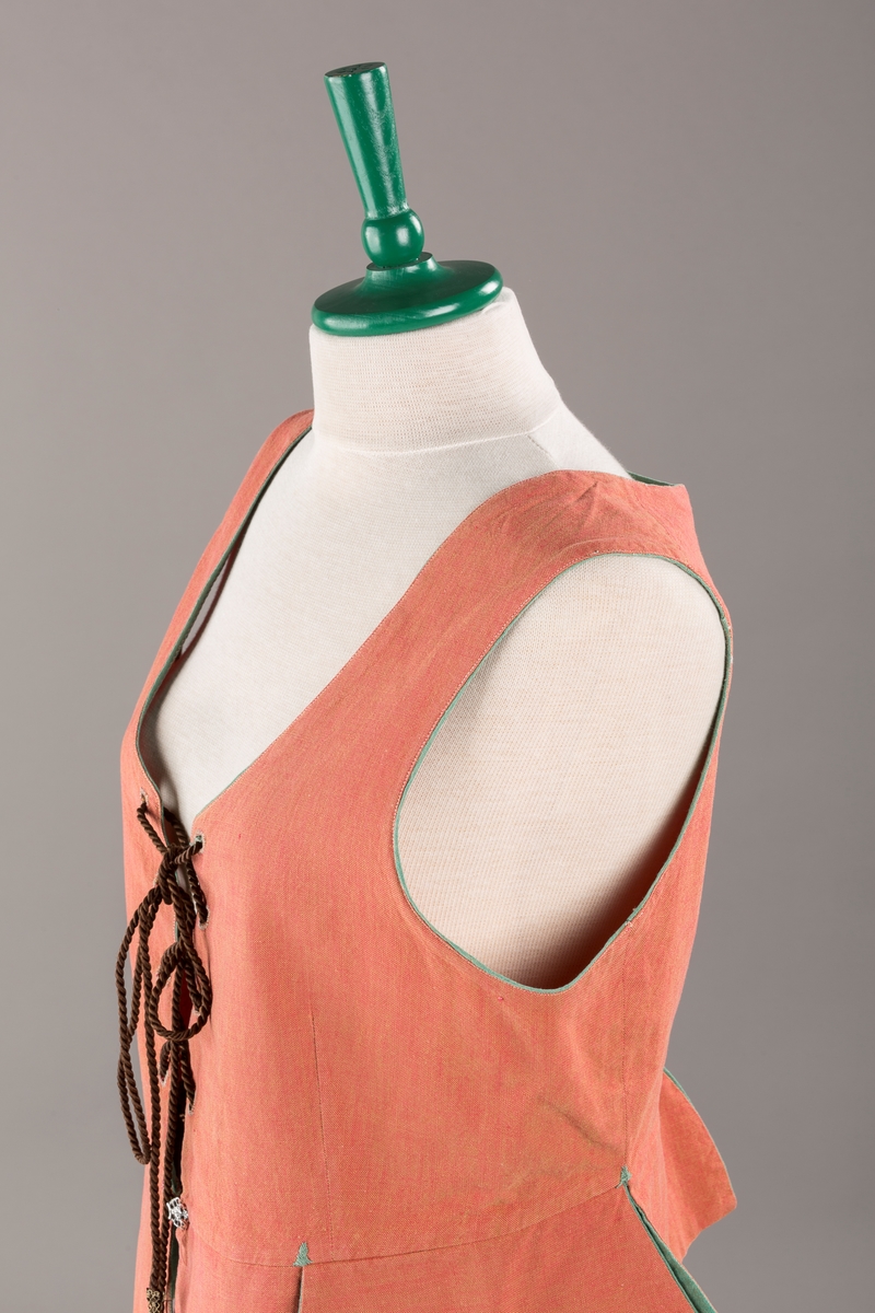 Rustrødt liv med grønn kant og fór, seks skjøter. Håndsydd forsterkning i kontrastfarge øverst i skjøtesplittene. Lukkes med tinnhekte og silkesnor.