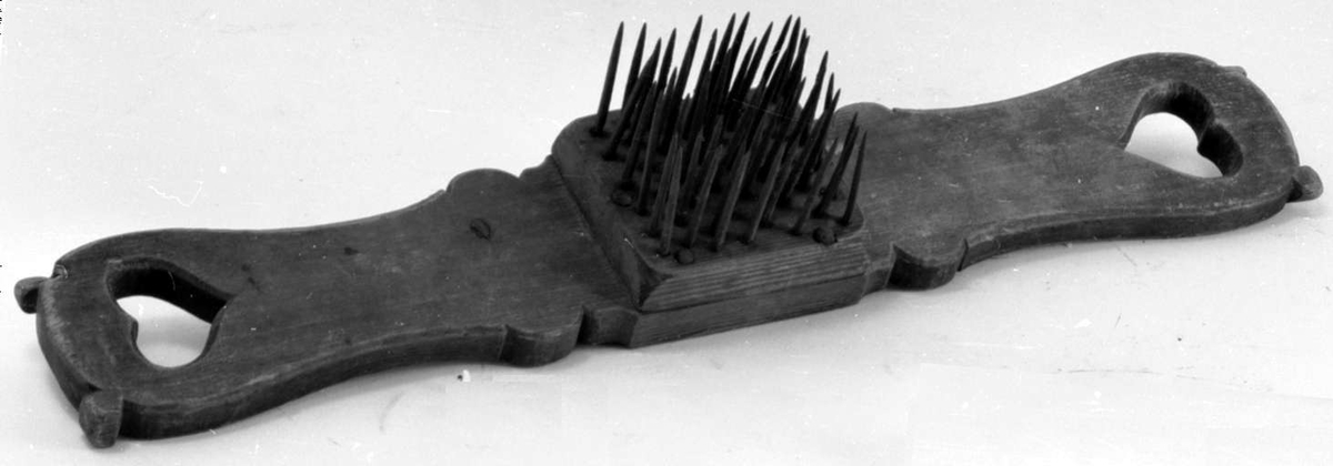 Linhekle av tre med hull som hjerter i hver ende av brettet. På midten er det festet en trekloss med stålspiker.