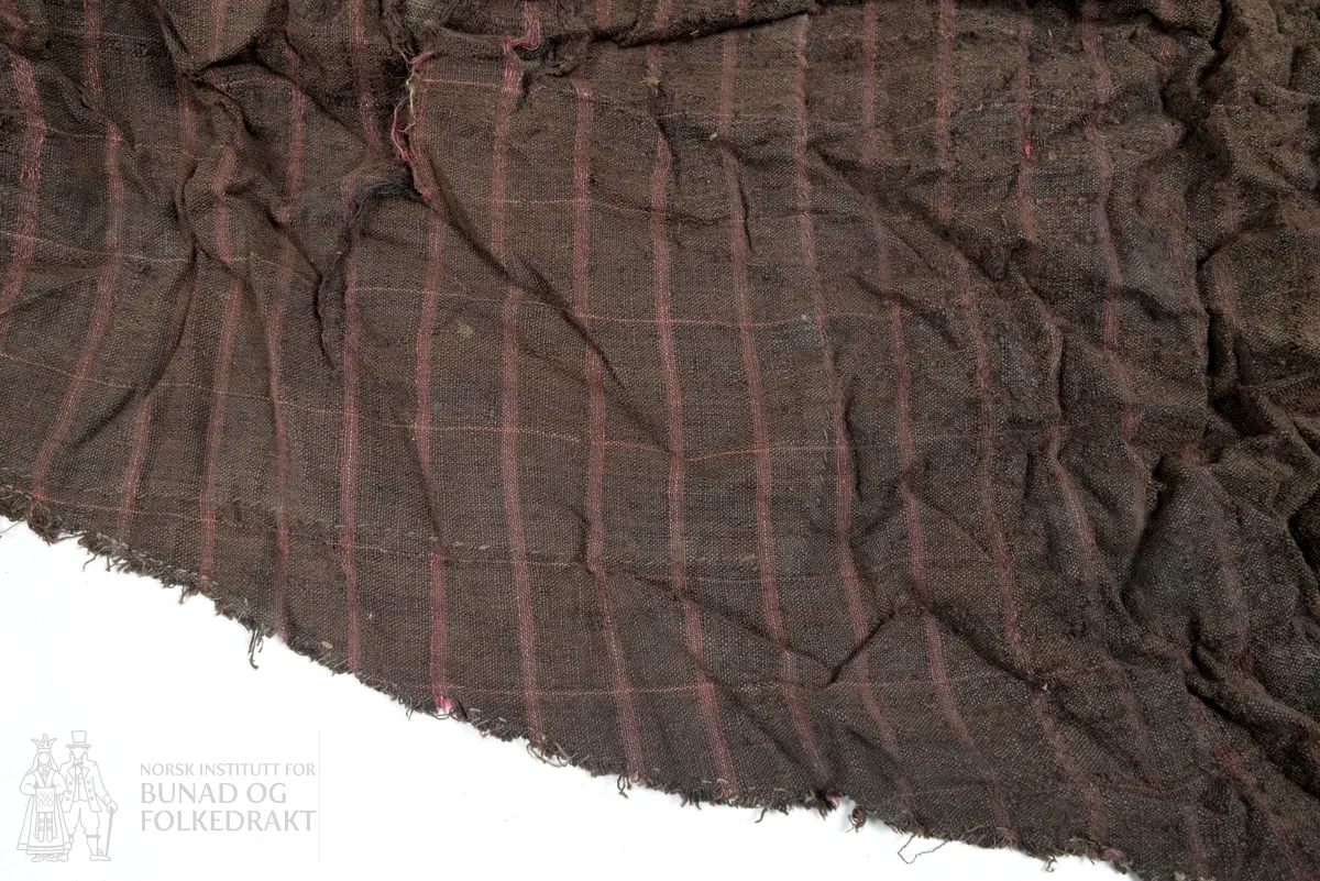 Svært utslitt del av ein stakk med linning, nederkant og ei metallhekte i eine enden av linning. Nedanfor denne delen av linning er det sauma fald som truleg er splitten midt framme på stakken. Av tre ulike verkensstoff, lerretsbinding med bomull i renning, ull i innslag. Stripete stoff i brun og lilla, svært utydeleg lillafargar. Fleire små hol etter mus, har vore nytta som stappefille.