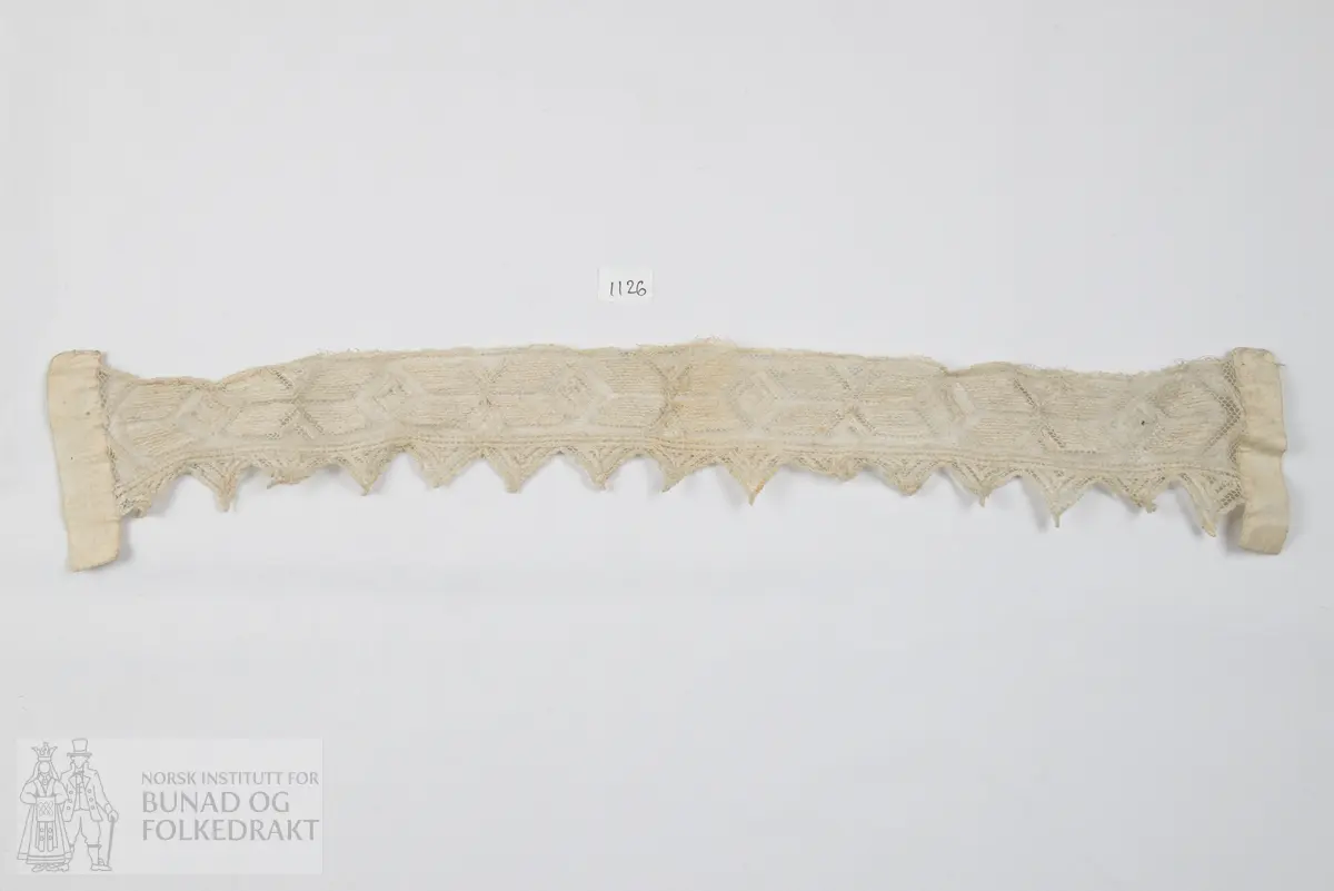 Ei lang remse av tyll forma med trekantsnippar etter eine langsida. Tyllstopping med grov laustvinna ujavn kvit lintråd - "gasisaum". I kvar ende er påsydd eit smalt dobbelbretta stoffstykkje av kvit linlerret.