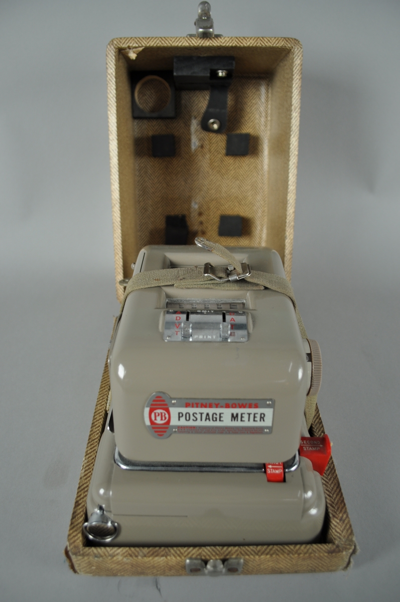 Frankeringsmaskin av brunt metall. 

Maskinen er plassert i trekasse med fiskebensmønstret utside. Kassen har håndtak.