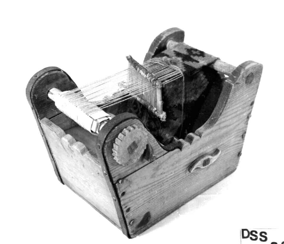 Trekasse med brystbom, garnbom og slagbom. Veven har renning og det er vevet et lite stykke bånd med mønster i rødt, blått og grønt.