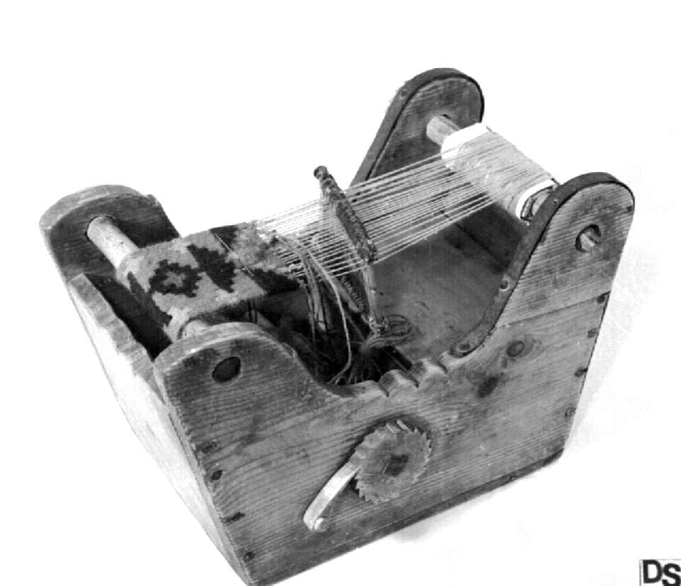 Trekasse med brystbom, garnbom og slagbom. Veven har renning og det er vevet et lite stykke bånd med mønster i rødt, blått og grønt.