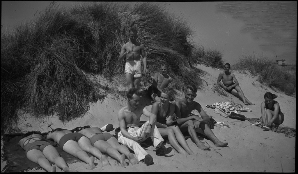 En stor vennegjeng i lystig lag på stranda på Sola. De har trommer og trekkspill og leker i vannet og på land med og uten ball. Sola strandhotell ligger i bakgrunnen. Tor Johannessen og Harald Bergsaker er på bilde nr. 14 og 18.