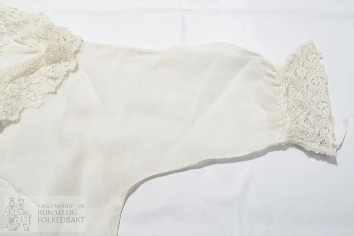 Dåpskjole i tynn hvit bomull, vevd i litt glissen lerretsbinding. Splitt midt bak, lukkes med knytebånd i nakken. Knytebåndet ligger i løpegang i halsen. Løpegang med knytebånd også nederst på ermene. Kragen er laget av kjøpeblonde/maskinprodusert blonde, samme kjøpeblonde er brukt som kanting nederst på skjørtet og nederst på ermene. Blondene er blitt stivet. Ikke tydelig definert liv, sydd med en lang sidesøm. Sidesømmen er sydd i ett med ermesømmen. Maskinsøm med håndsydde detaljer.