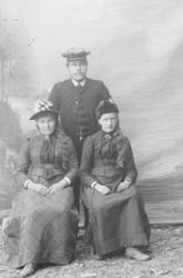 Mann står bak to kvinner som sitter på stol. Nevnt Margith J