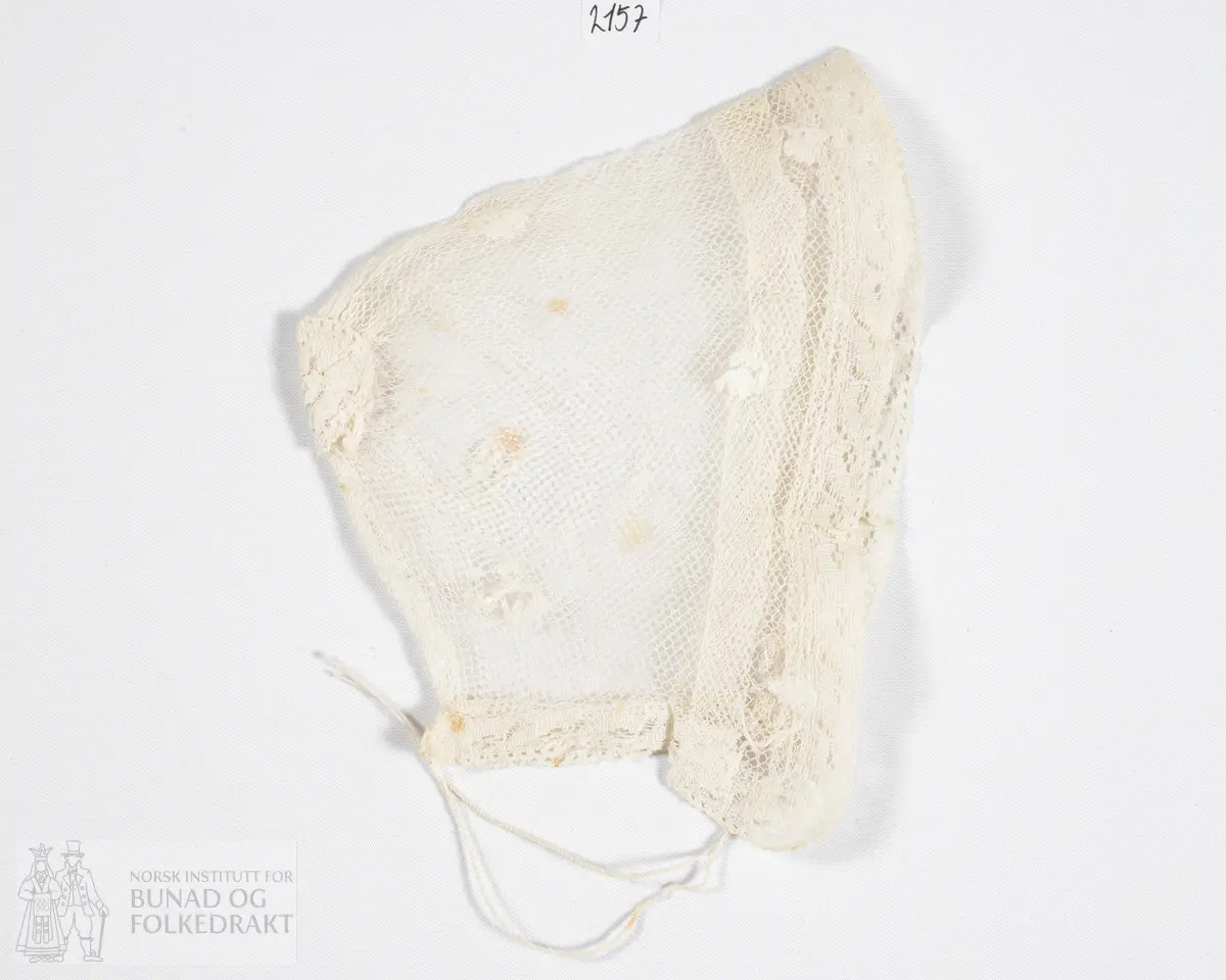 Barnelue i stivet hvit bomullstyll. Kantet med kjøpeblonde. Brodert med små blomster fordelt rundt på luen, broderiene er gjort i bomullsgarn. Smalt tvunnet knytebånd i bomull, ligger i løpegang nederst på luen.