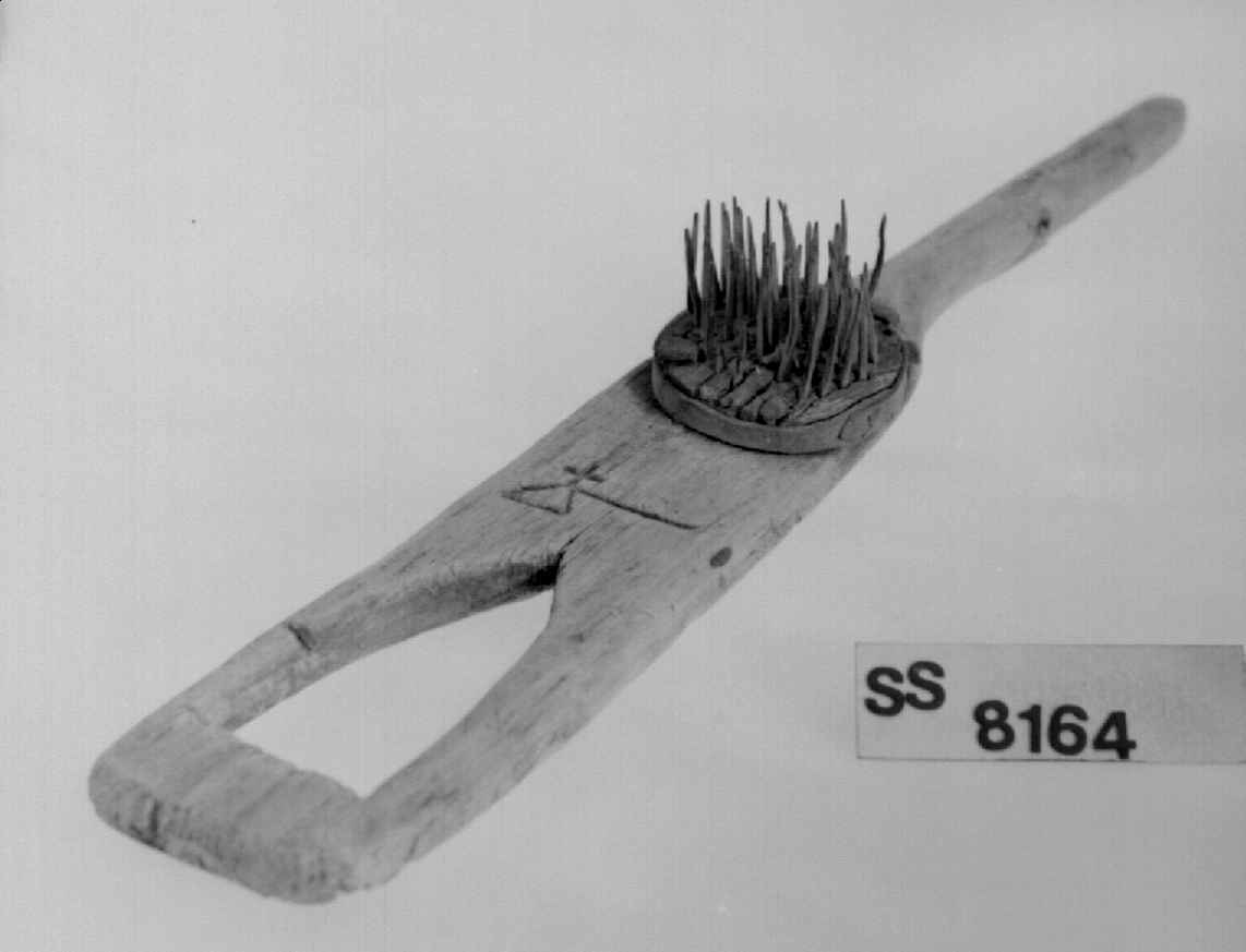 Linhekle som har et håndtak i den ene enden, den andre enden er smal og spiss. På midten av heklen er det en rundt kloss med stålspiker. Det er skåret ut et merke på brettet bestående av en trekant, en strek og et kors.