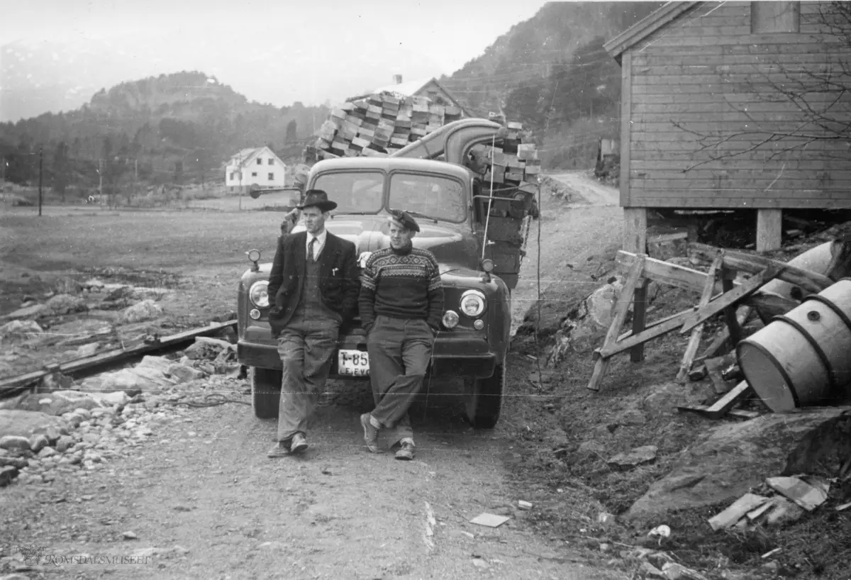 Lastebilen til Jofred Kirkeslett (1932-2000), med et lass med plank på vei ner i Heggdalsviken. Ragnar Heggdalsvik (1926 - 1978) står ved siden av Jofred. Ragnar omkom ved uhell i skogen..Huset til vendtre tilhører Harald Orset. Huset knapt synlig bak lastebilen er butikken til "Per i vikjen"