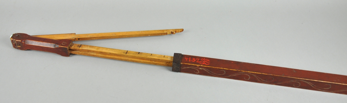 Målestokk av tre der tommestokken - som har to ledd - trekkes ut av den ytterste hylsen. Hylsen er dekorert med en bord / ranke. Kanten på hylsen er forsterket med metall.