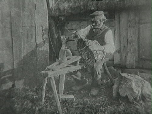 Henrik Bengtsson i södra Björsgård sitter på en 'träskomärr' - en anordning man använde vid träskotillverkning - och tillverkar en korg. Han har en pipa i munnen, träskor och sitter vid en skiftesverkslada med halmtak. (Se även bildnr VMA 6719_2.)