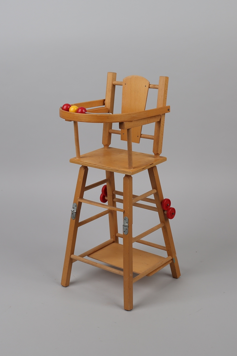 Dukkestol, laget som barnestol med bord foran setet. bena på bord og stol er hengslet. Bordet kan svinges under stolbena slik at stolen blir høy. De to fremste bordbena og de to bakerste stolbena har hjul, slik at når stolen er i lav posisjon, kan den trilles. 3 plastkuler montert på metallstang foran på spisebrett.