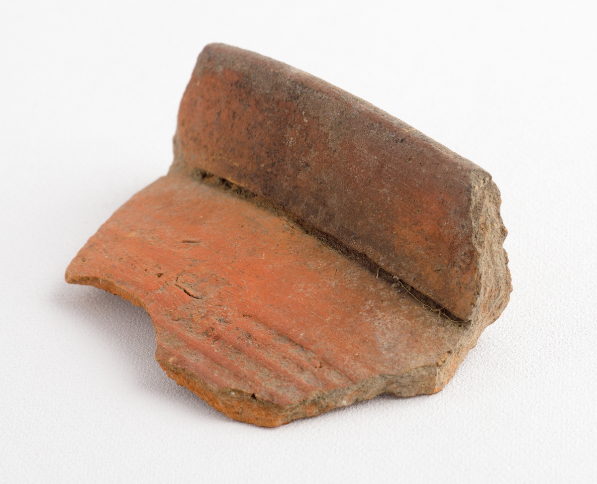 Fragment av mynning till ett keramikkärl, möjligen en trefotsgryta. Mynningen är av yngre rödgods och har en utkragad kant, samt utvändiga drejlinjer. Brun glasyr på kärlets insida. Tillverkad i södra Skandinavien.