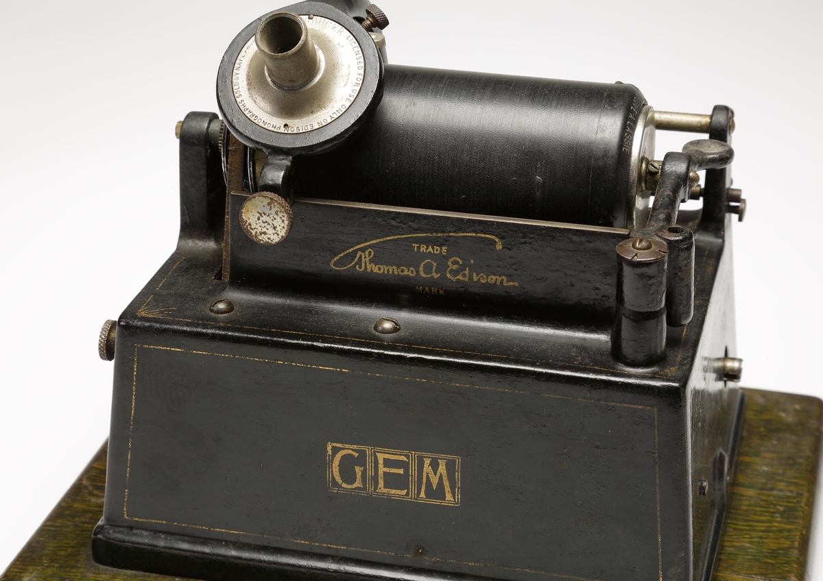 Fonograf, med förvaringslåda. 
Rektangulär, svartlackerad metallåda med musikinstrumentet ovanpå. Dessutom en förnicklad (?) tratt. Vrides upp med nyckel på kortsidan. 
Förvaringslåda av fernissat trä med handtag och påmålad text: "Edison, GEM, Phonograph".