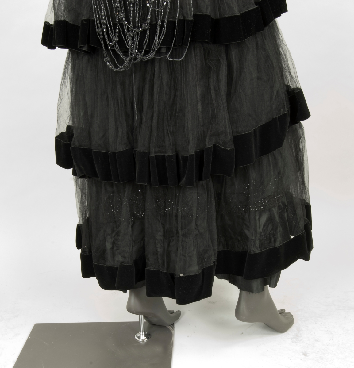 Klänning av svart tyll. Livet broderat med pärlor och paljetter. Ankellång kjol av volanger kantade med svarta sammetsband. Fyrkantig halslinning och kort ärm med stor vidd. Baktill dekorerad från axlarna med långa pärl- och paljettband. 1910-tal.