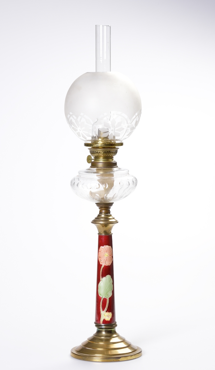 En st. bordslampa/fotogenlampa av majolika. 
Hög modell med blomdekor i jugend mot rödbrun bakgrund. Lamphus av glas med olivslipning. Rund skärm av glas med matterad dekor. Rund, terrasserad fot av mässing. Elektrifierad (?).