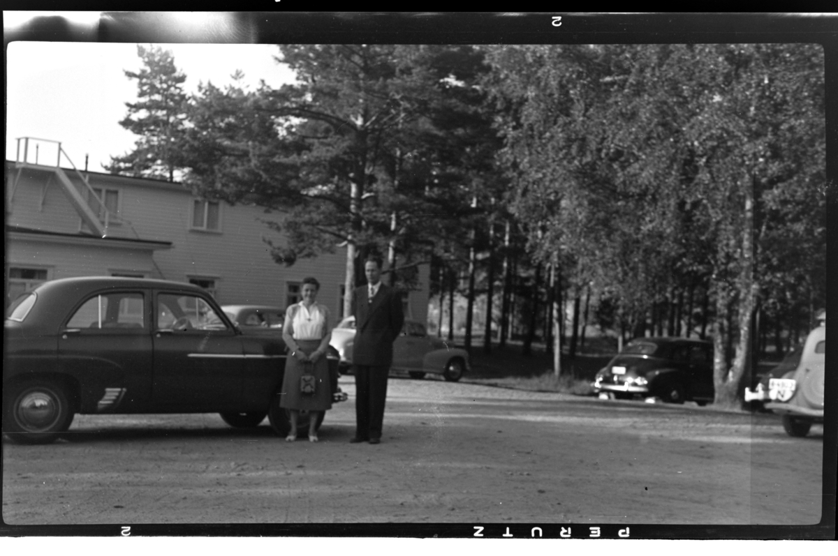 Et ukjent par står foran en bil. Fotografert ca. 1953.