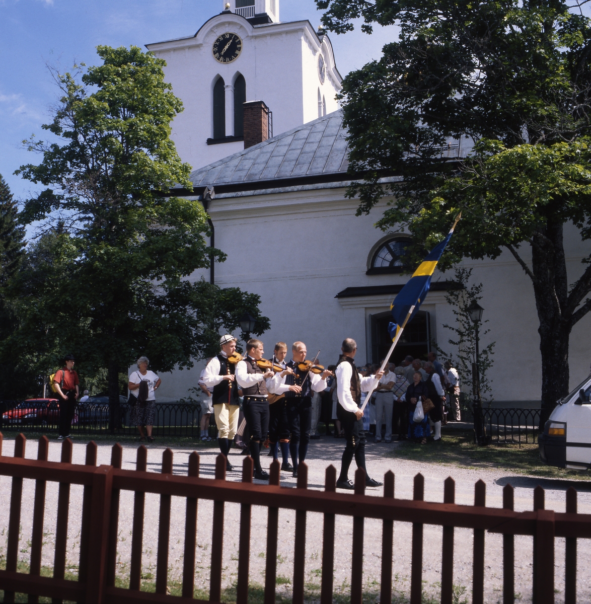 Rengsjöfesten 11 juli 1999. Här står musiker i folkdräkt och en man med fana utanför kyrkan.