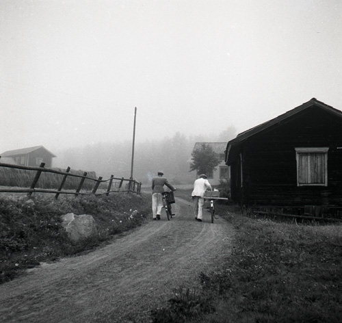 Två män leder sina cyklar på en grusväg mot några gårdar i dimma, Knåda, Edsbyn september 1949.