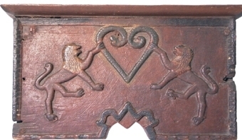 Handdukshängare av trä målad i brunt och grönt. På framsidan i relief två lejon med varsin tass placerad på en hjärtformig mittfigur.