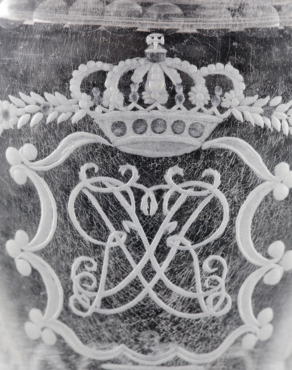 Pokal av glas, med lock. 
Slipat och graverat mönster med kungligt spegelmonogram: "FR"* i kartusch under krona. Invikt kant på foten.

*"FR" (Fredericus Rex= Fredrik I av Sverige, reg. 1720-1751).