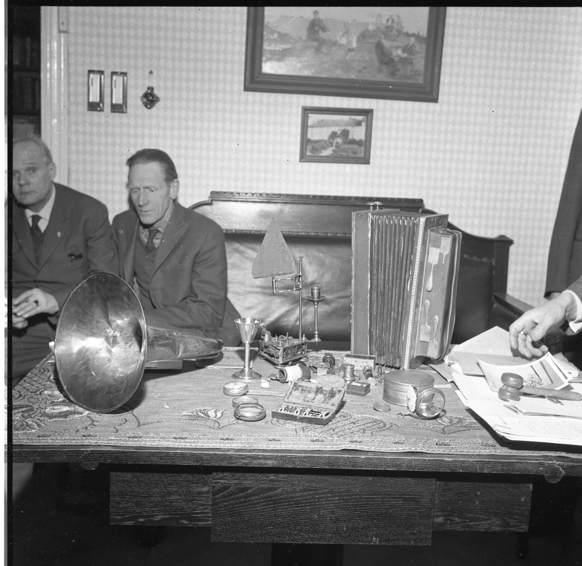Holger Andersson och Bernt Rendahl sittande i vänsterkanten bakom ett bord med olika föremål: tuba, skospännen, dragspel mm. Sannolikt nyförvärv till Grenna Hembygdsförening. Somliga skänkta av Åke Barck Berkén.
