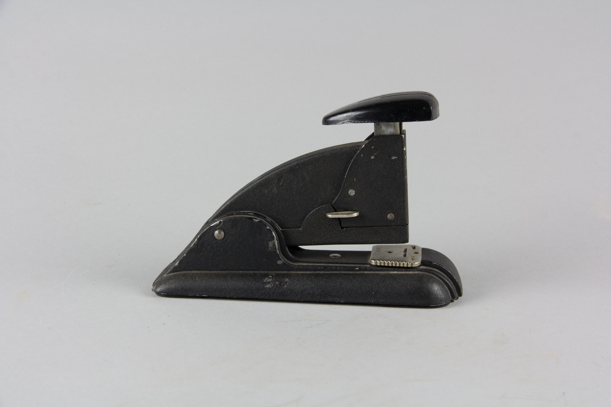 Häftapparat Speed Stapler tillverkad av svartlackerad metall. Avlång fotplatta med överdel. Tryckhandtag i plastmaterial högst upp. Innehåller ett fåtal häftklamrar.