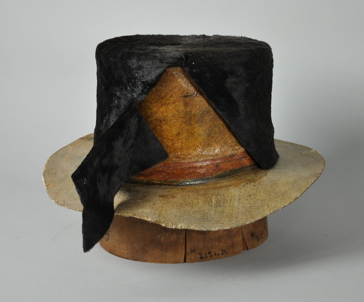 Hatteblokk med påsatt hatt av strie. Selve hatteblokken er satt sammen av fem deler av tre, som sammen utgjør en sylinderform med innfelt midtparti. Hatten av strie har brem, og på pullen er det satt på uferdig parti med skinn.