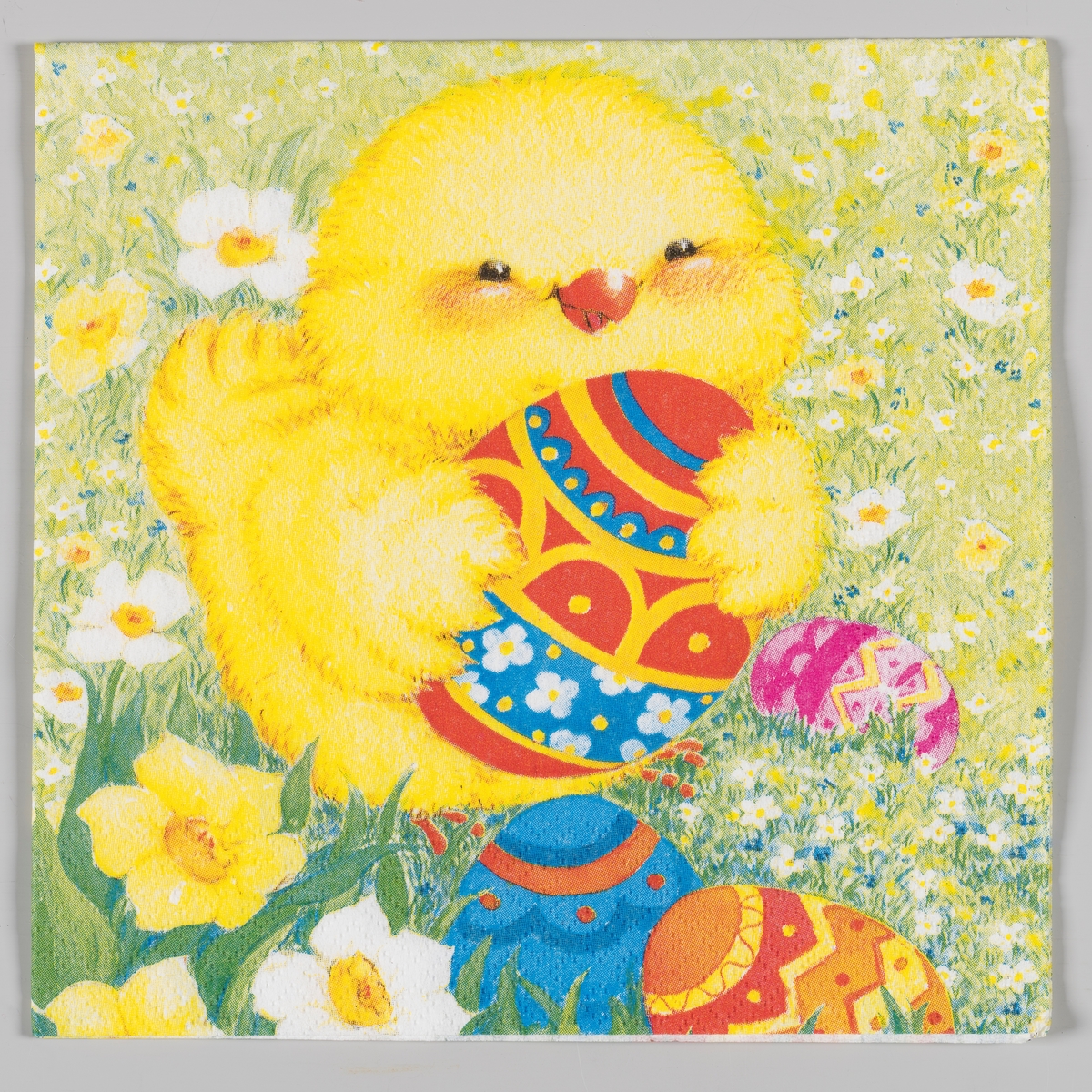 En kylling holder et dekorert påskeegg med vingene sine. Dekorerte påskeegg, påskeliljer og hvite og blå blomster i gresset rundt kyllingen.