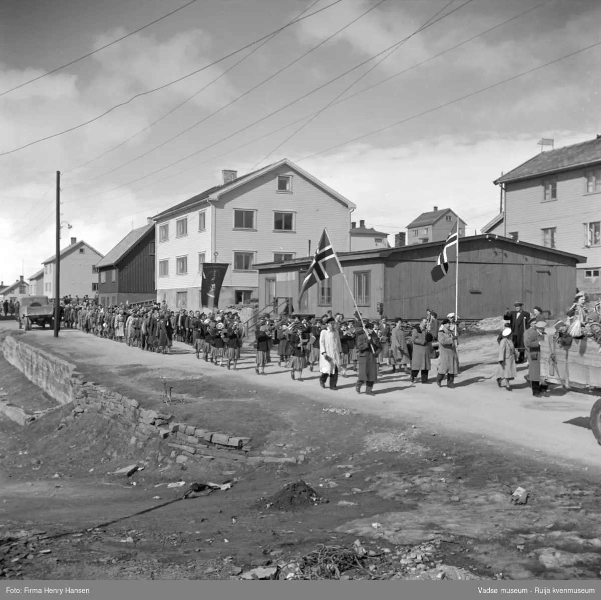 Vadsø 17 mai 1952. 17.maitog på vei østover i Havnegata, med muskkorps og fane. Det står en trehjulssykkel i forkant av bildet.