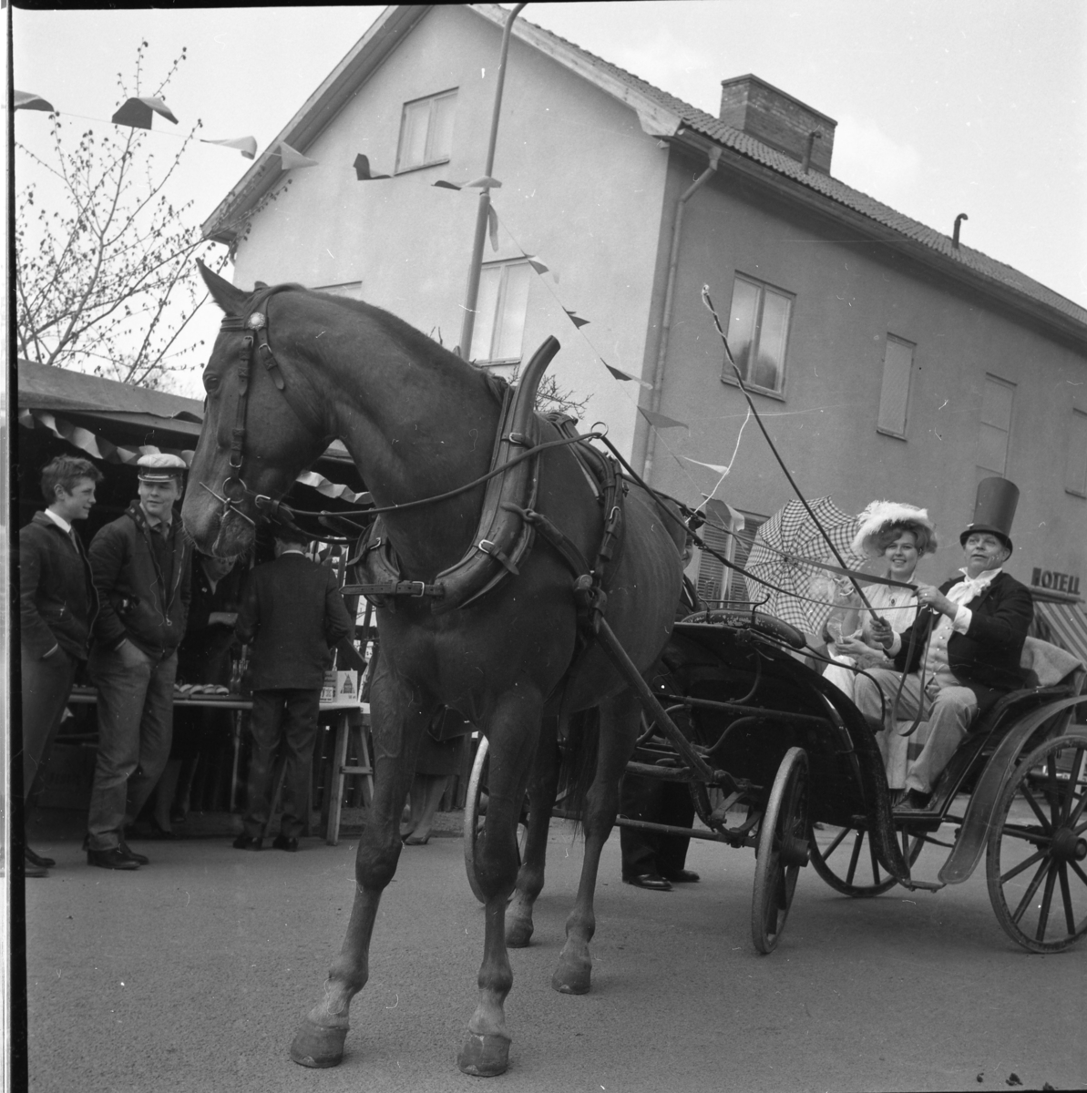 Lions vårmarknad 1964. En högtidsklädd Lauritz Madsen med extra hög hatt sitter i hästvagn och håller tömmarna. Intill sitter dottern Anne-Lise Madsen med hatt och parasoll. På Sjögatan vid korsningen mot Ribbagårdsgränd. På Sjögatan 29 var det då ett Hotell.