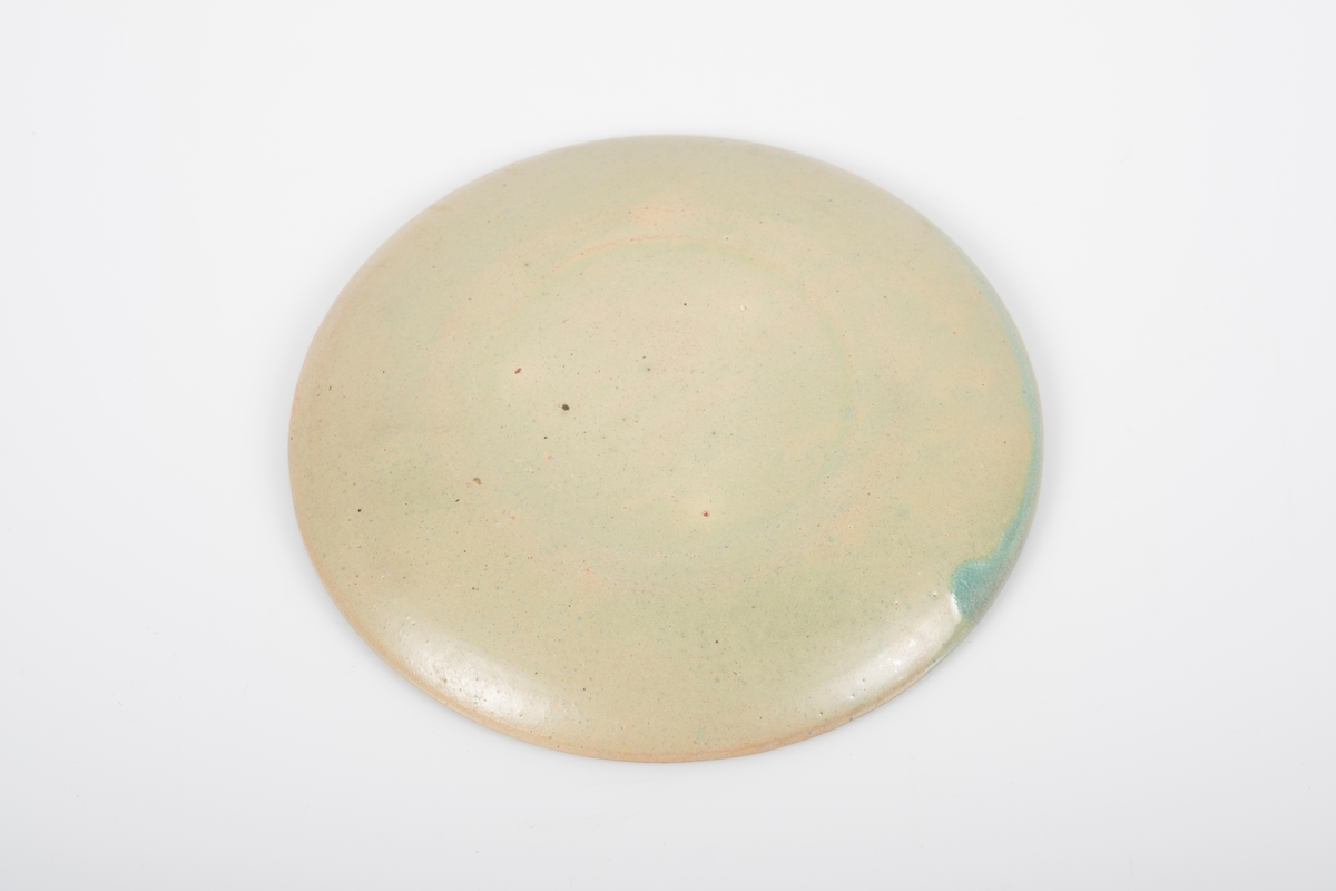 Rund skål i keramikk med grønn lasur. Buet hank på koppen. Skålen er blank på oversiden og matt på bunnen. Det mangler lasur på to små område.