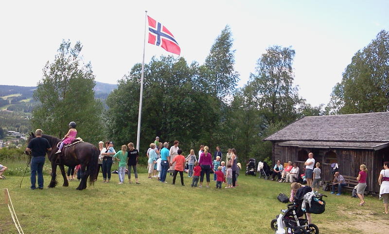 Arrangement på tunet. Flagget er heist, og flere personer har kommet for å hygge seg. Her er det også en hest med en unge på ryggen.