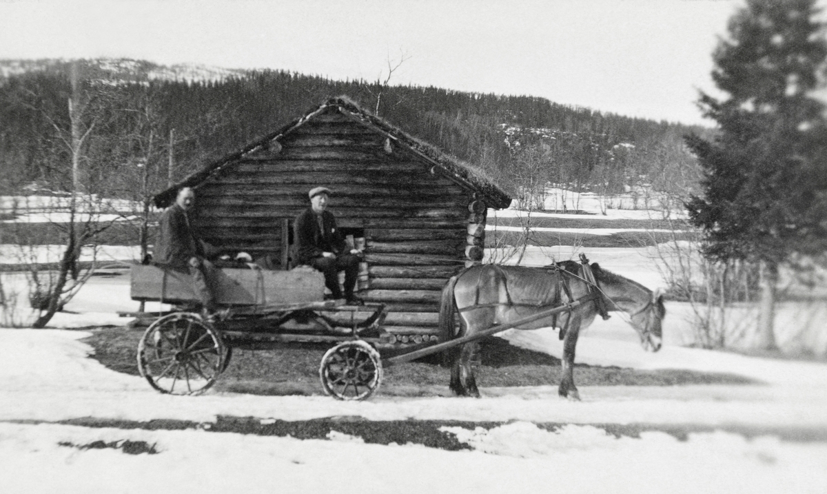 Hest med vogn og to passasjerer.  Fotografiet er tatt i føreløsningen i et landskap som er flekkevis snødekt.  Hesten og kjøretøyet er oppstilt utenfor gavlen på ei høyløe.  I bakgrunnen skimtes engesletter. 