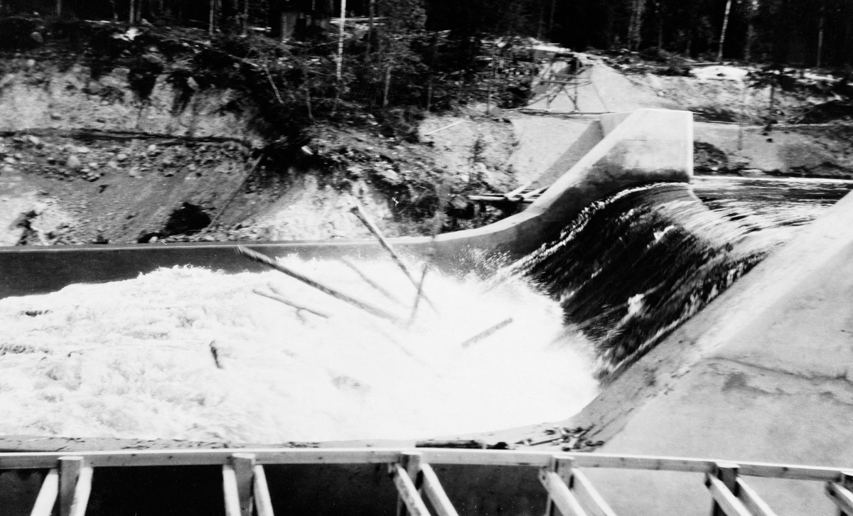 «Nydammen» ved Osfallet i elva Søre Osa i Åmot i Hedmark, fotografert da denne dammen var nybygd, sommeren 1917.  Denne dammen ble reist som erstatning for den opprinnelige kraftverksdammen på dette stedet, som ble fullstendig ødelagt under flom i mai 1916, bare to år etter at den ble satt i drift (jfr. fanen «Opplysninger»).  Den avbildete dammen var langt mindre og et enklere provisorium, bygd for å få i gang igjen eletrisitetsforsyninga til bygdesenteret Rena og den treforedlingsbedriften som var under etablering der. Fotografiet viser en armert betongdam med kraftige sidevanger. Vi ser også hvordan vannmassene, etter å ha passert damterskelen, falt ned i et kvitskummende strømkav der tømmerstokkene kunne reise seg på høykant.  Dette fenomenet var et tegn på at tømmeret var utsatt for store påkjenninger når det passerte dammen, noe som neppe var popluært i Kristiania Tømmerdirektion, som administrerte fløtinga i dette sidevassdraget til Glomma.