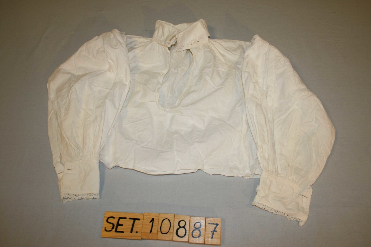 Kvit bomullsskjorte i tradisjonell form. Lange ermar, blonder på krage og mansjettar. Sliten