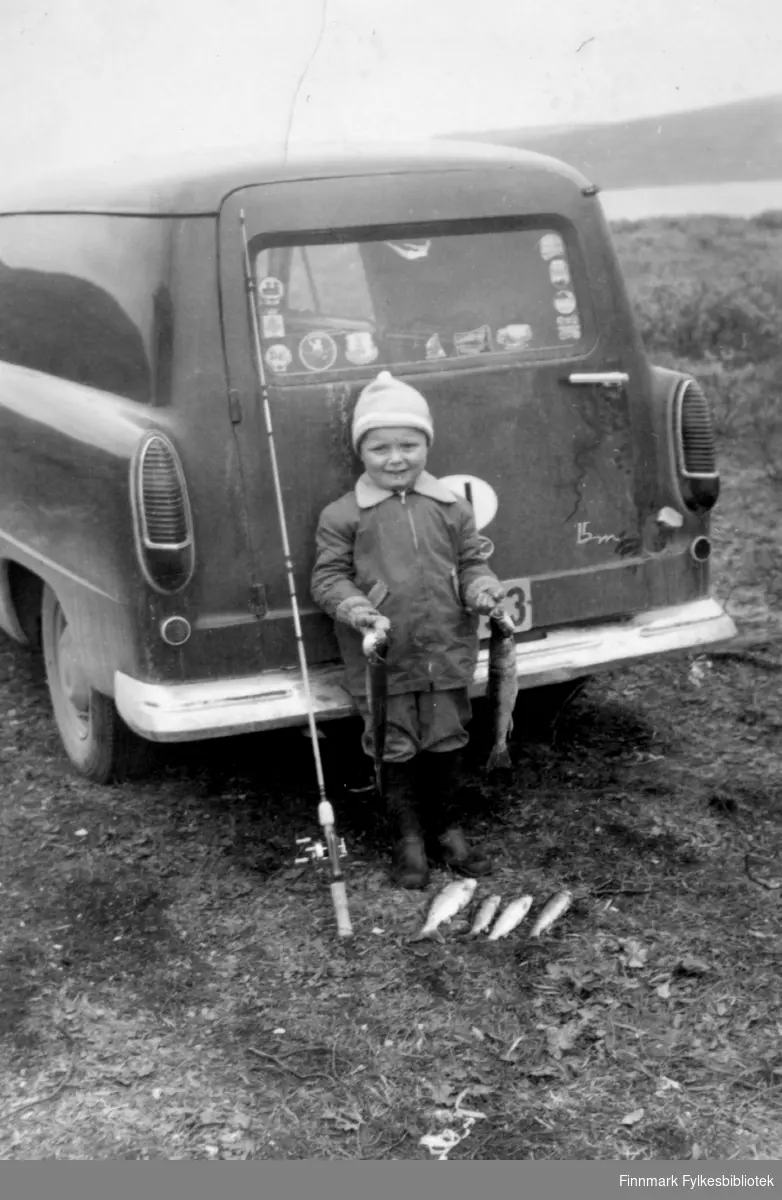 Barn med fiskefangst fotografert bak en Ford Taunus 15M varebil. Kfr 15M-logo står nederst til høyre bak på døren. Barnet står med fiskestang og flere fine ferskvannsfisker liggende på bakken. Barnet holder en fisk i hånden. Langs kanten av bilruta er det klistrert på mange merker med stedsnavn. Det var vanlig å kjøpe slike på bensinstasjoner når man var ute på ferieturer. Bildet kan være tatt i perioden 1955-1970. 

Familiealbum tilhørende familien Klemetsen. Utlånt av Trygve Klemetsen. Periode: 1930-1960.
